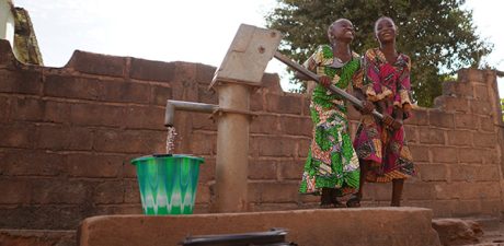 Two girls in colorful clothing pump water from a hand well into a bucket, smiling as they work together in a rural community. Photo © CIVIC